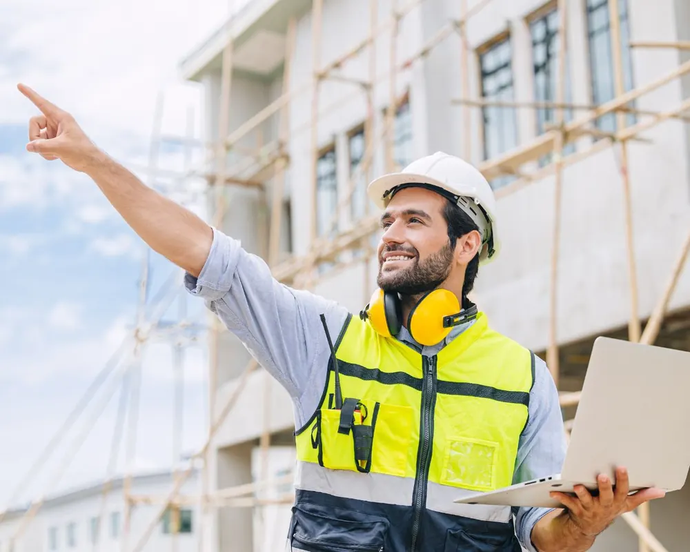 Encadrant en casque blanc et gilet haute visibilité qui indique une direction sur un chantier, un ordinateur portable à la main